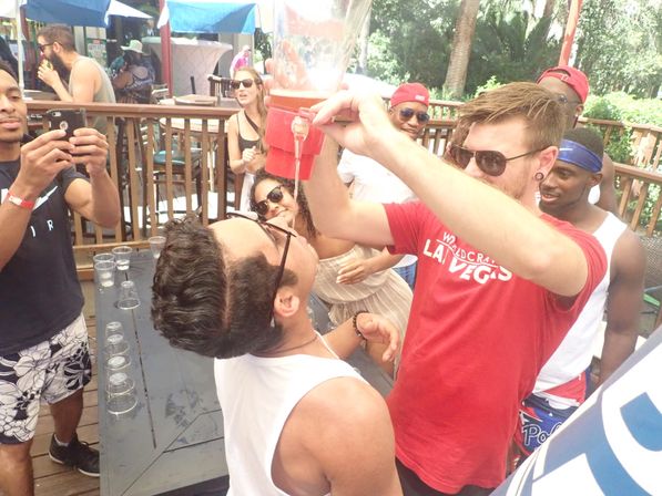 Outdoor bar patio party with friends doing a beer funnel from a red drink tower, people cheering and taking photos in sunglasses