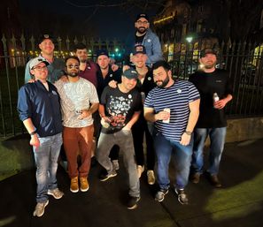 Group of eleven men on a city sidewalk at night by a wrought-iron fence, enjoying a night out with drinks under street lamps