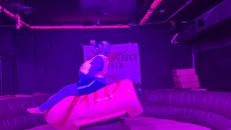 Person in blue outfit riding a novelty mechanical hot dog on an inflatable ring under purple stage lights in an indoor nightlife venue