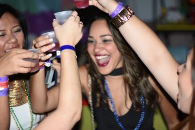 Friends cheering and clinking plastic cups, wearing colorful wristbands and bead necklaces at a lively indoor party.
