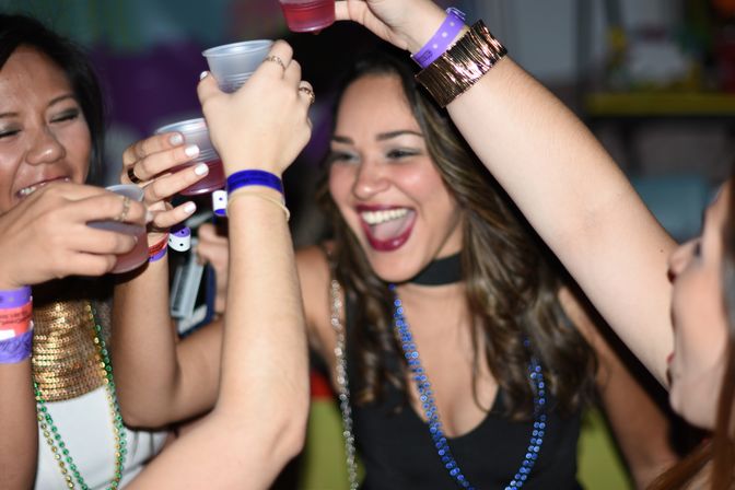 Friends cheering and clinking plastic cups, wearing colorful wristbands and bead necklaces at a lively indoor party.