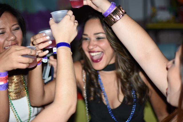 Friends cheering and clinking plastic cups, wearing colorful wristbands and bead necklaces at a lively indoor party.