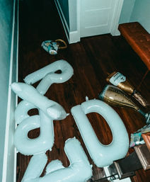 Party aftermath in a home hallway: deflated white inflatable number balloons and gold champagne-bottle foil balloons scattered on dark hardwood floor beside a tufted bench and closed white door.