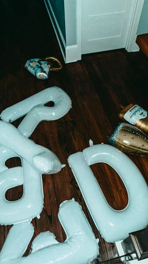 Party aftermath in a home hallway: deflated white inflatable number balloons and gold champagne-bottle foil balloons scattered on dark hardwood floor beside a tufted bench and closed white door.