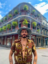 Traveler in a wide-brim hat and patterned shirt on a New Orleans French Quarter street, framed by a historic building with ornate wrought-iron balconies and hanging ferns.