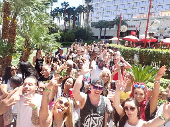 Crowd of smiling people at a sunny outdoor hotel pool party surrounded by palm trees, red umbrellas and high-rise windows, many holding drinks and taking selfies.