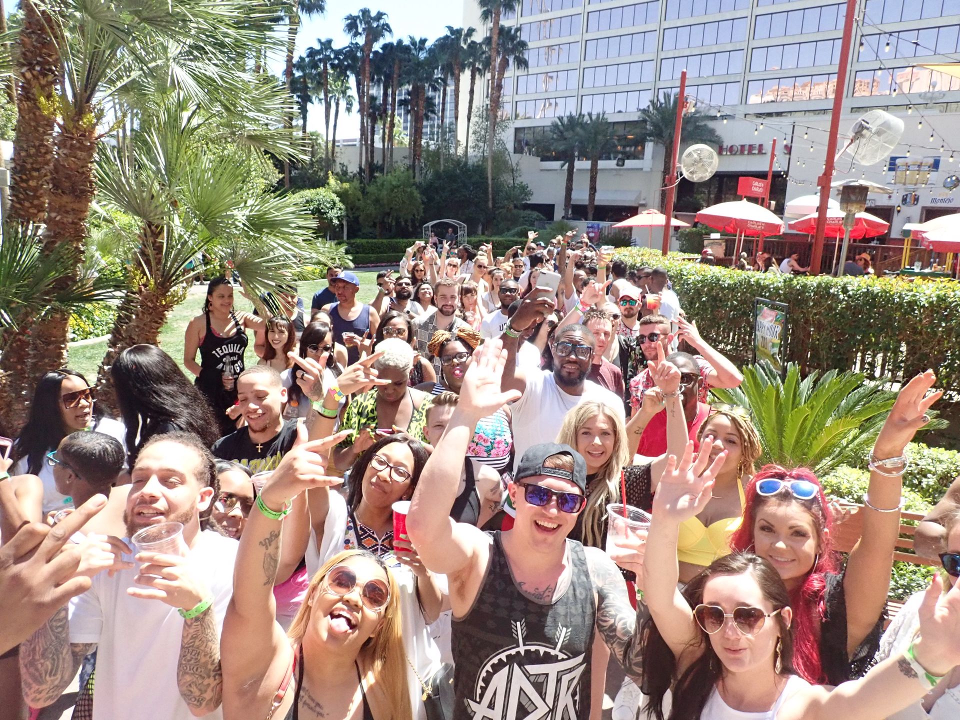 Crowd of smiling people at a sunny outdoor hotel pool party surrounded by palm trees, red umbrellas and high-rise windows, many holding drinks and taking selfies.