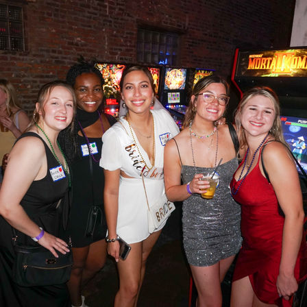 Bachelorette party at an arcade bar — five friends smiling for a night out, bride-to-be in white sash and purse, pinball and arcade machines behind them