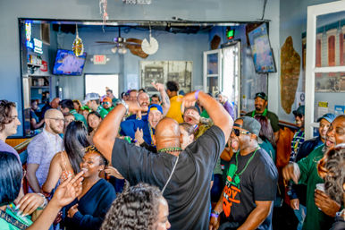 Lively crowd of adults packed into a neighborhood bar, dancing, laughing, and wearing green beads during a festive daytime celebration