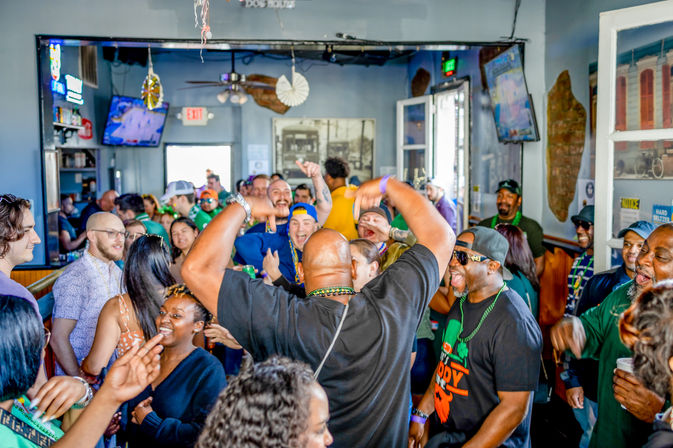 Lively crowd of adults packed into a neighborhood bar, dancing, laughing, and wearing green beads during a festive daytime celebration