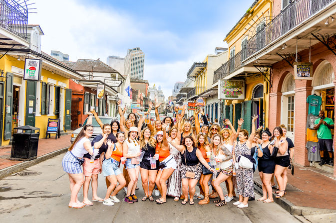Cheerful large group of women posing and celebrating on a lively New Orleans French Quarter street lined with colorful storefronts and wrought-iron balconies under a bright sky