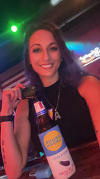 Smiling woman at a neon-lit bar holding a beer wrapped in a bright blue-and-yellow koozie, American flag and neon sign in the background