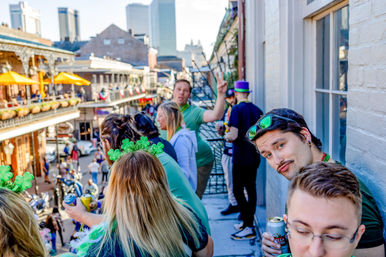 Group of people in green on a New Orleans French Quarter balcony watching a lively street celebration, wearing shamrock accessories and holding drinks