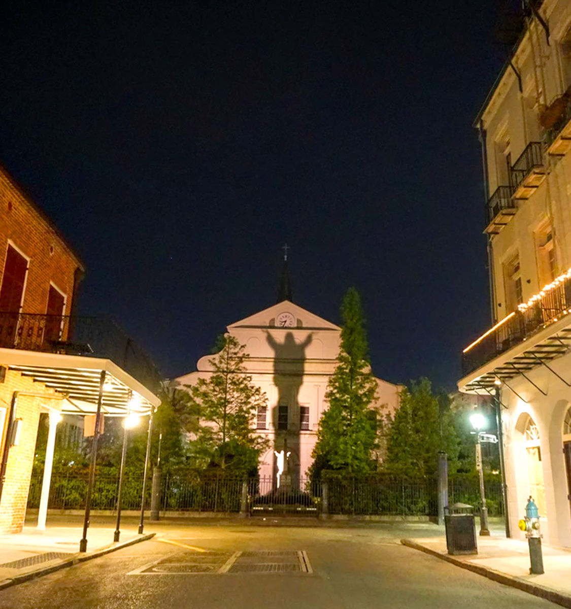 Night scene in New Orleans French Quarter: cathedral façade illuminated with a giant statue shadow, flanked by balconies, trees, and street lamps.