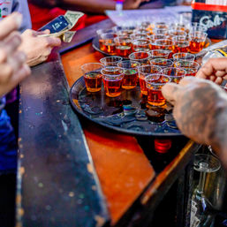 Tray of amber liquor shots in plastic cups on a busy bar counter, a tattooed server handing out drinks while patrons exchange cash — lively nightlife bar scene.
