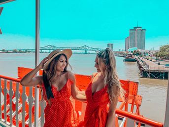 Two women in red polka-dot summer dresses smiling on a riverboat deck with an orange railing, one wearing a straw hat, with a bridge and waterfront skyline in the sunny background.