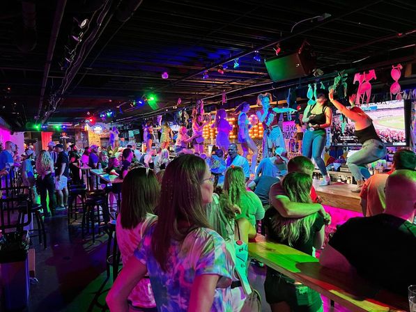 Lively neon-lit bar scene with a crowded crowd watching performers dancing on the bar counter under colorful stage lights and bras strung above, energetic city nightlife vibe