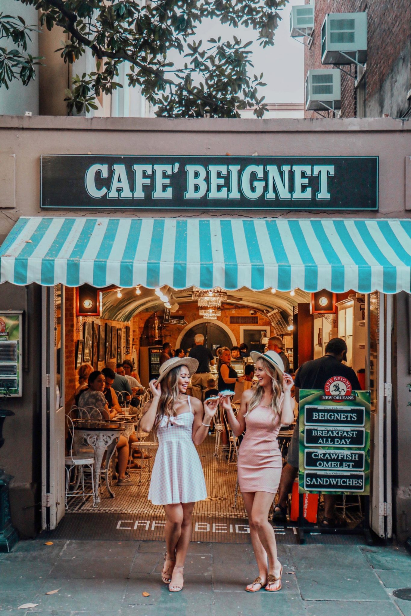 Two women in sun hats and summer dresses laugh while sharing beignets outside a café with a turquoise-striped awning and cozy indoor seating visible.