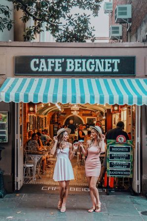 Two women in sun hats and summer dresses laugh while sharing beignets outside a café with a turquoise-striped awning and cozy indoor seating visible.