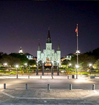 Night view of St. Louis Cathedral and Jackson Square in New Orleans, featuring a lit equestrian statue, rows of streetlamps, and an American flag