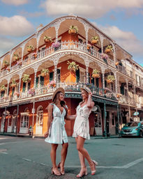 Two friends in sundresses and sun hats pose on a street in front of a colorful ornate wrought-iron balcony building with hanging plants in the French Quarter, New Orleans.