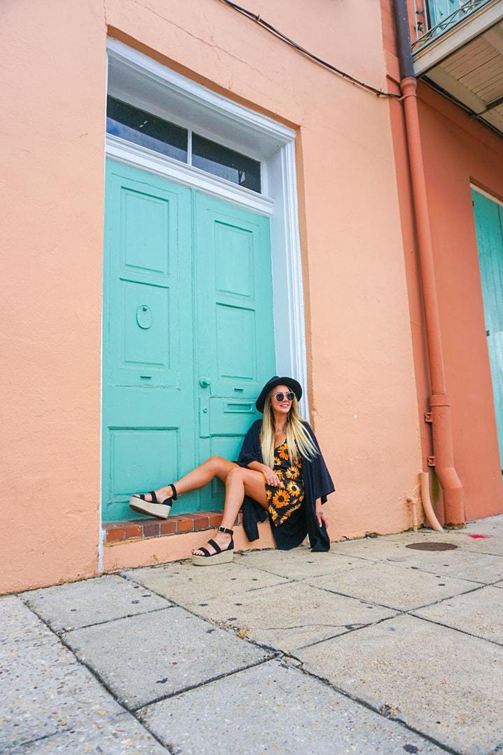 Fashionable woman in a sunflower-print dress, black hat and sunglasses sitting on the step of a teal door set in a peach pastel building — vibrant street-style scene on an urban sidewalk.