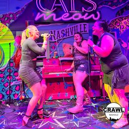 Three singers performing karaoke on a neon-lit Nashville stage with a pink piano, star-patterned floor, colorful leopard-print backdrop and bright magenta and purple lighting.
