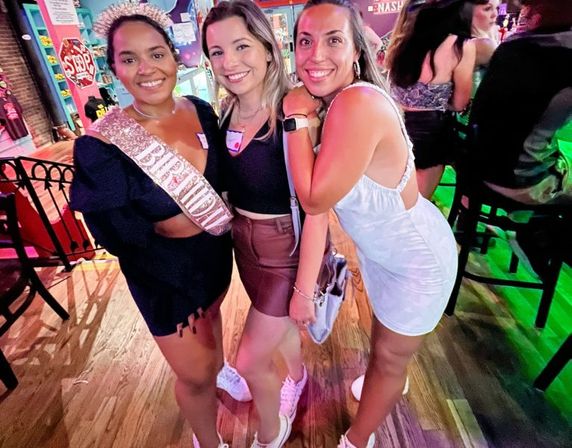 Three smiling friends posing inside a lively bar for a night out; one wearing a birthday sash and tiara, casual dresses, wooden floor and colorful neon lighting.