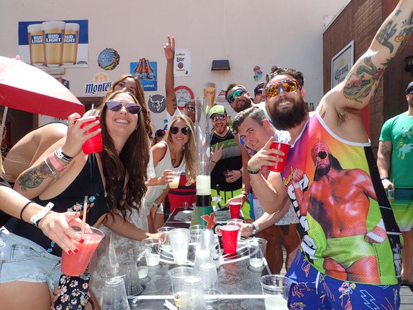 Sunny outdoor patio party with a group of friends smiling and raising red cups around a tall drink tower, wearing sunglasses and tattoos against a backdrop of beer signs.