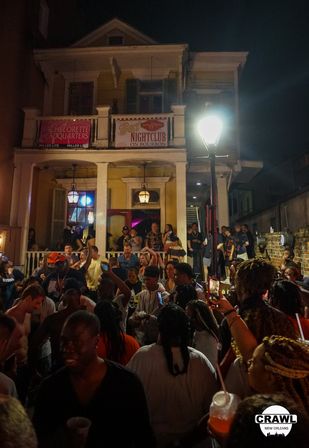 Lively New Orleans French Quarter nightlife scene: crowded street party outside a two-story balcony bar with banners, a bright streetlamp, and people holding drinks at night.