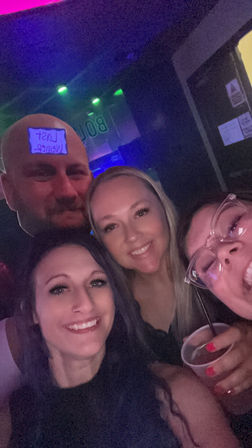Nightclub selfie of four friends smiling under neon lights, holding drinks and laughing — fun night out vibe