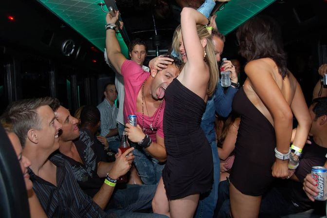Crowded party bus interior at night with a lively group of young adults dancing and drinking; a man in a pink shirt playfully sticks out his tongue near a woman in a black dress while others cheer and hold cans and bottles.