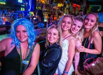 Five friends smiling at a neon-lit bar for a night out — one in a bright blue wig with beads, others holding drinks with bartenders and bottles in the colorful background.