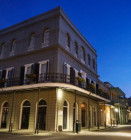 Twilight view of a historic three-story corner building with arched windows, black shutters, and an ornate wrought-iron balcony above an empty street.