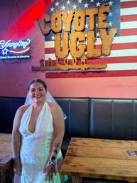 Smiling bride in a white halter wedding gown and veil poses inside an American-themed saloon bar by a large rustic wooden sign on a flag mural, neon beer sign and reclaimed-wood tables.