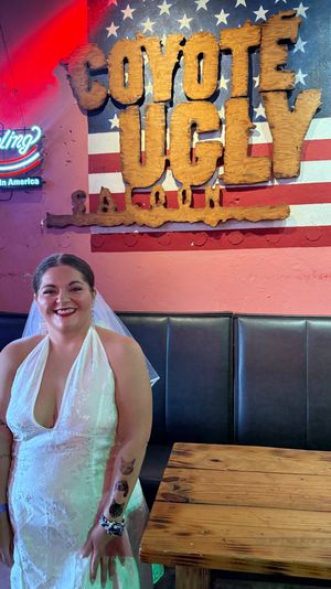 Smiling bride in a white halter wedding gown and veil poses inside an American-themed saloon bar by a large rustic wooden sign on a flag mural, neon beer sign and reclaimed-wood tables.