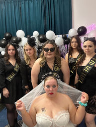 Indoor bachelorette party group portrait: bride in white lace corset and veil seated in front of bridesmaids dressed in black with sashes, veils and heart sunglasses, with black-and-white balloon and metallic fringe backdrop.