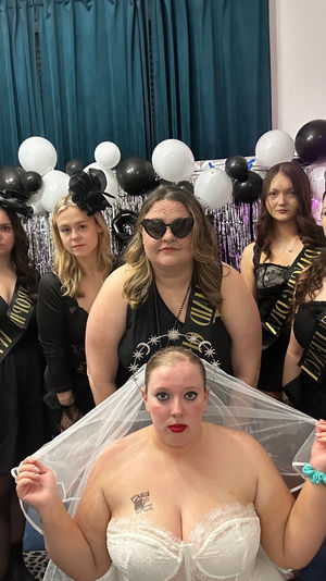 Indoor bachelorette party group portrait: bride in white lace corset and veil seated in front of bridesmaids dressed in black with sashes, veils and heart sunglasses, with black-and-white balloon and metallic fringe backdrop.