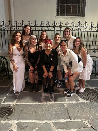 Nine women cheerfully posing on a stone sidewalk at night in front of a decorative iron fence — group celebration in an urban historic district.