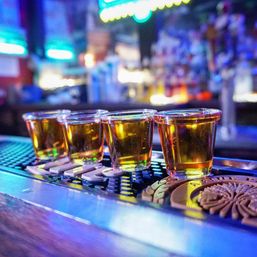 Four golden whiskey shots in plastic shot glasses lined up on a bar mat on a neon-lit counter, colorful bokeh background evoking lively nightlife and downtown bar scene.