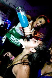 Bartender in a green apron playfully pouring a bright blue cocktail from a large bottle into a woman’s open mouth at a crowded nightclub bar with neon lights and party vibes.