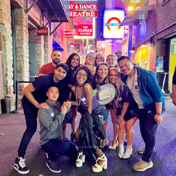 Smiling group of friends posing in a lively neon-lit downtown nightlife alley at night, colorful signs and marquees overhead and party outfits and accessories creating a fun urban entertainment vibe.