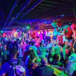 Vibrant neon-lit bar packed with revelers dancing on the bar and at tables under colorful stage lights — lively downtown nightlife scene.