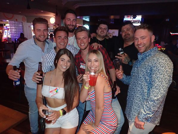 Smiling group of friends posing with beers and cocktails in a dimly lit bar with neon lights — lively nightlife group photo.