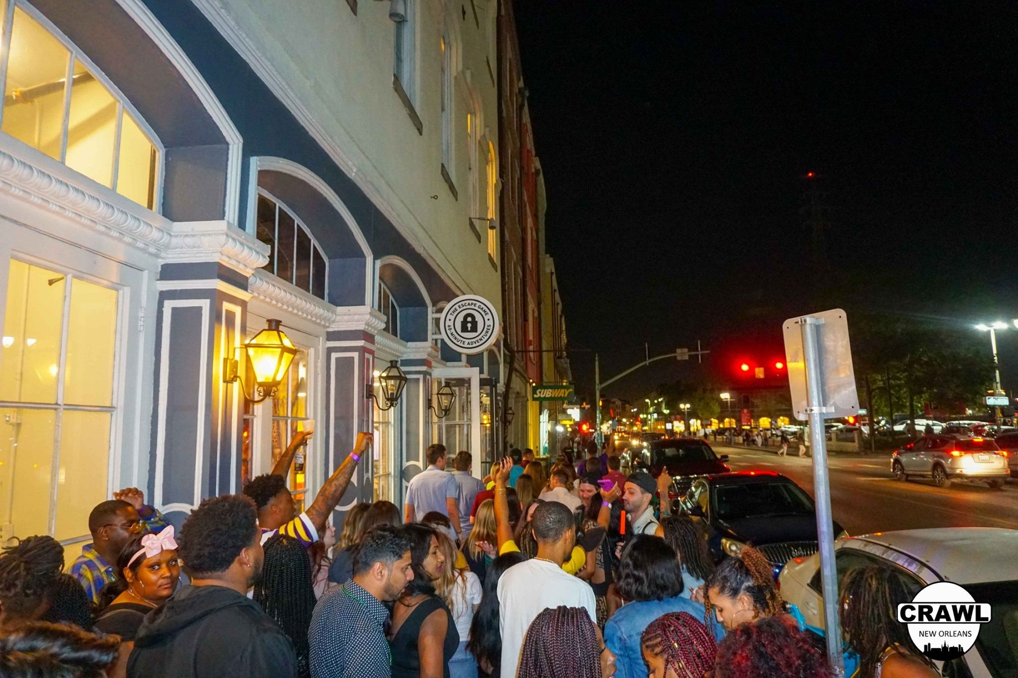 Lively nighttime downtown crowd queued along a lamp-lit historic storefront with arched windows, parked cars and traffic lights on a busy city street — urban nightlife scene.