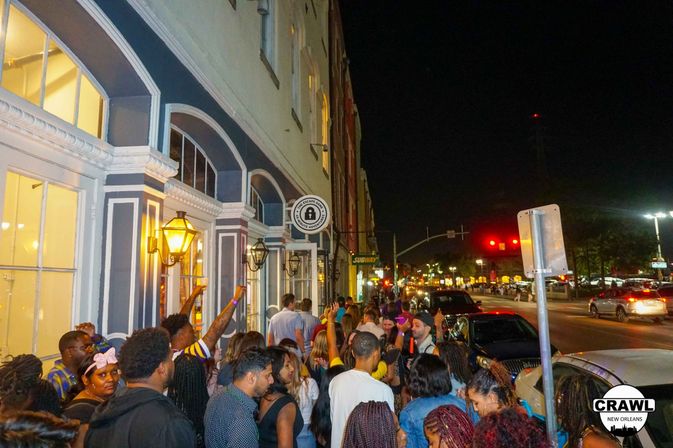 Lively nighttime downtown crowd queued along a lamp-lit historic storefront with arched windows, parked cars and traffic lights on a busy city street — urban nightlife scene.