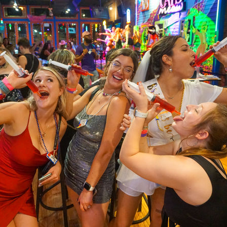Bachelorette party friends in a neon-lit bar taking red cocktail shots from syringe-style dispensers, laughing and celebrating a night out