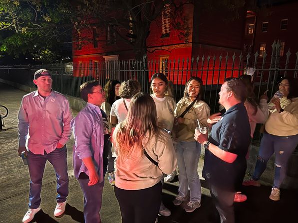 Late-night urban scene: group of friends chatting and laughing on a city sidewalk with drinks, beneath trees by a wrought-iron fence and red-lit brick building.