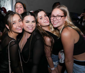 Five young women smiling and posing together at a crowded indoor nightclub/party, one holding a canned drink — casual black tops and denim, city nightlife vibe.
