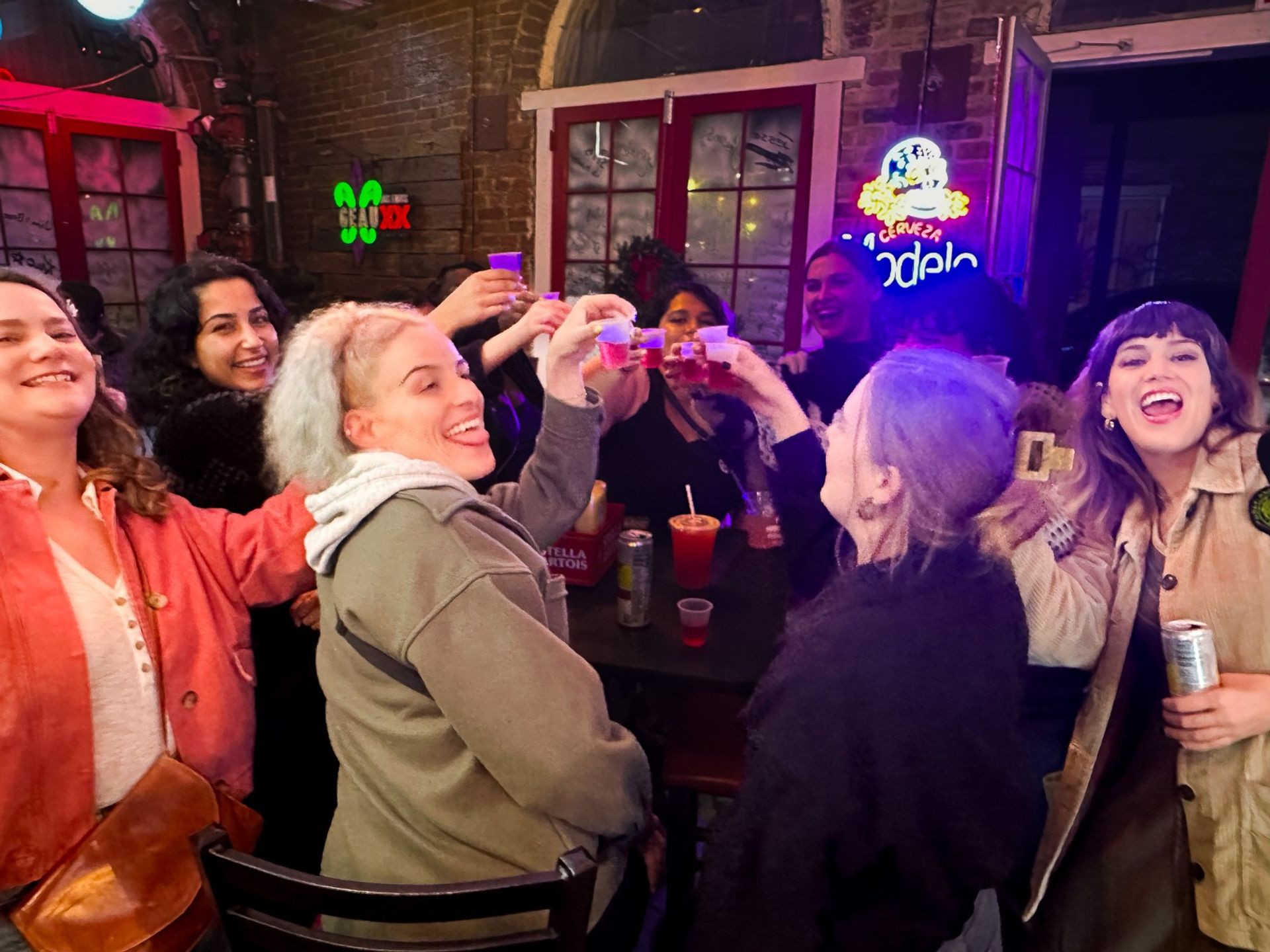 Lively group of friends cheering with plastic shot cups at a neon-lit downtown bar, brick wall and windows in the background — upbeat nightlife scene.